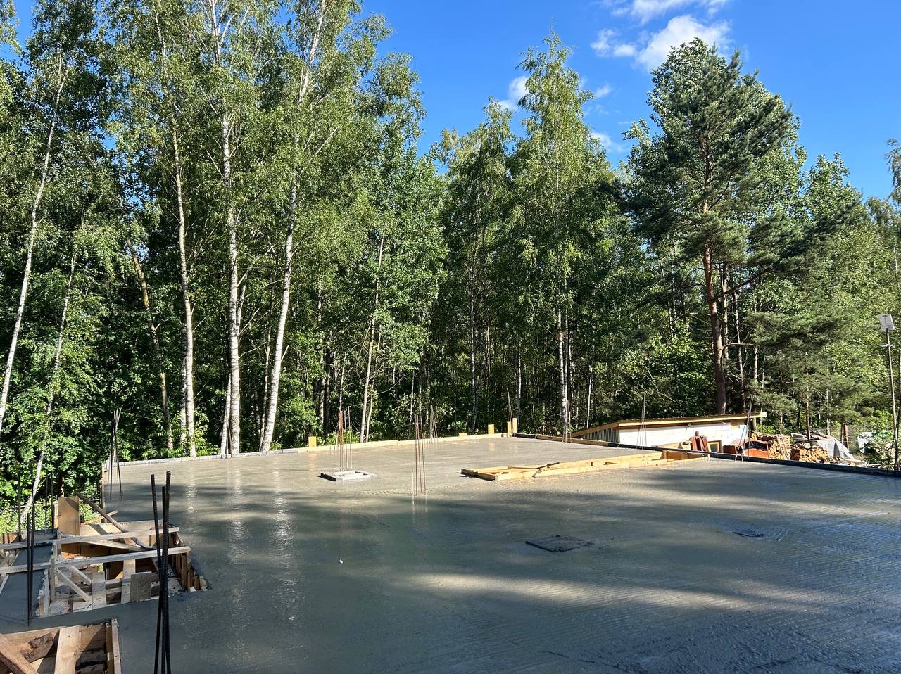 Wylany, świeży beton na płycie fundamentowej domu w otoczeniu lasu, widoczne wystające pręty zbrojeniowe i szalunki.