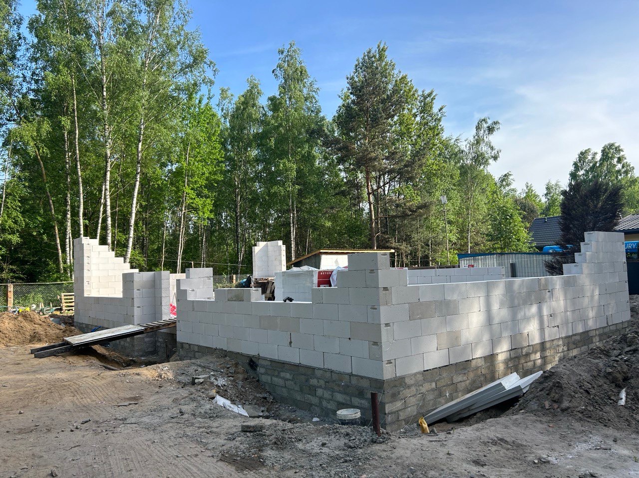 Wznoszenie ścian parteru z białych bloczków betonu komórkowego na fundamencie z cegły, widok na plac budowy otoczony drzewami w słoneczny dzień.