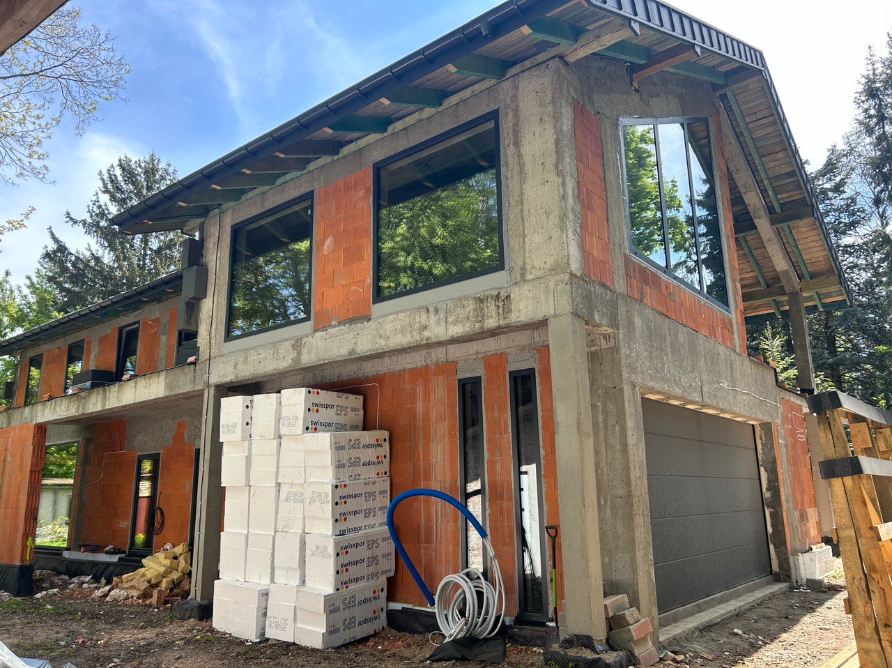 Dwukondygnacyjny budynek w stanie surowym otwartym z zamontowanymi oknami, widoczne bloczki ceramiczne, betonowe nadproża, stos białych płyt izolacyjnych i zwinięty wąż instalacyjny.