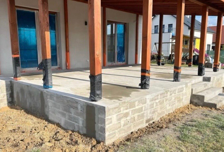 Wzniesiony fundament pod taras domu z drewnianymi słupami zabezpieczonymi folią, widoczne schody wejściowe i częściowo zagospodarowany teren.