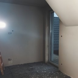 Kam-Art - Poddasze w trakcie prac wykończeniowych: ściany przygotowane do malowania, okno dachowe, zabezpieczona podłoga, widoczne kable oświetleniowe i drabina.