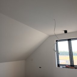 Kam-Art - Poddasze po świeżym malowaniu na biało, z widocznym oknem w czarnej ramie, wystającym kablem ze sufitu i narzędziami na podłodze.