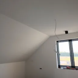 Poddasze po świeżym malowaniu na biało, z widocznym oknem w czarnej ramie, wystającym kablem ze sufitu i narzędziami na podłodze.
