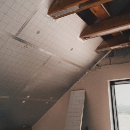 Kam-Art - Poddasze w trakcie ocieplania: widoczne drewniane belki stropowe, płyty izolacyjne z nadrukowaną siatką, częściowo wykończona ściana szczytowa z oknem.