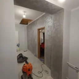 Korytarz w trakcie prac wykończeniowych: ściany pokryte dekoracyjną szarą fakturą, widoczny odkurzacz przemysłowy, zakryte meble w garderobie i niedokończony sufit z odsłoniętą konstrukcją.