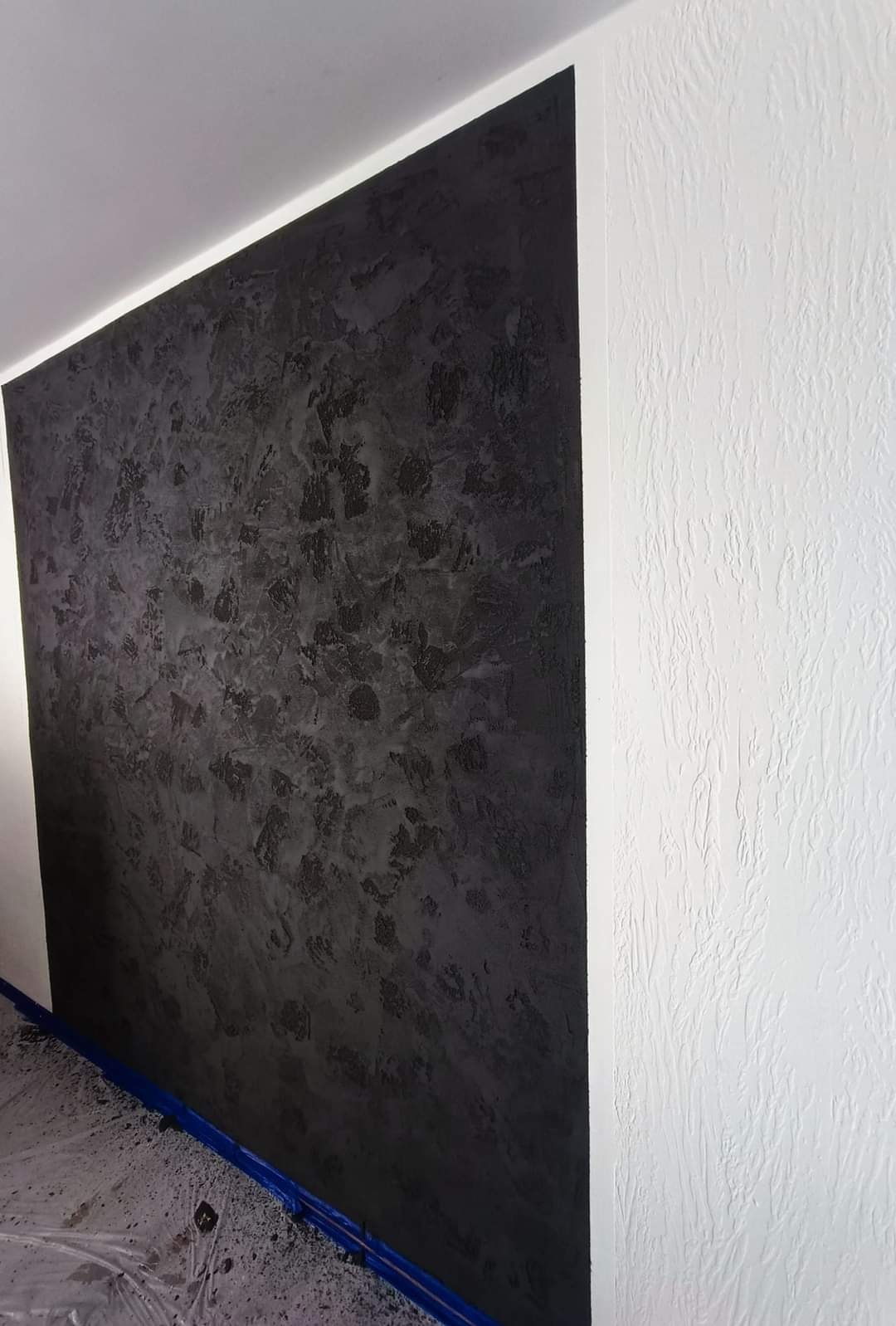 Częściowo wykończona ściana z dekoracyjną, ciemnoszarą fakturą obok białej ściany z wyraźną strukturą, zabezpieczona folią podłoga.