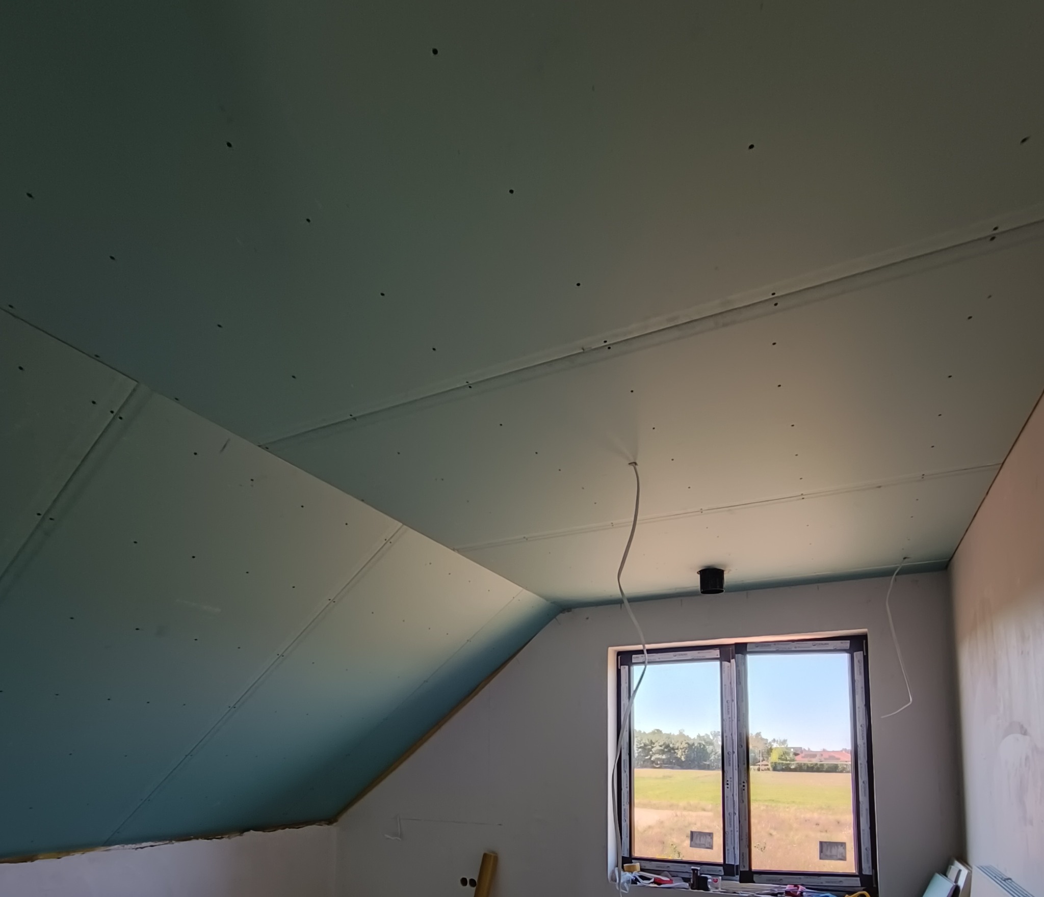 Poddasze z zamontowaną płytą kartonowo-gipsową przed szpachlowaniem, widoczne wkręty i wystające przewody elektryczne, okno z widokiem na zewnątrz.