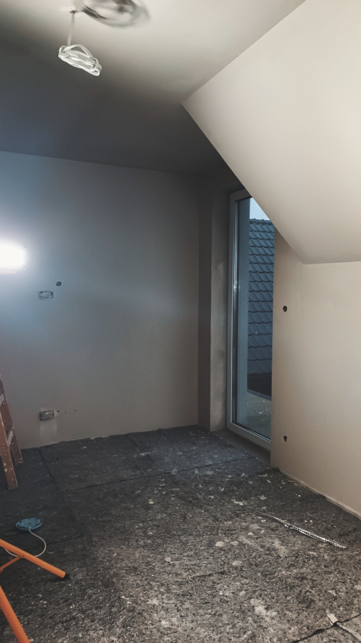 Poddasze w trakcie prac wykończeniowych: ściany przygotowane do malowania, okno dachowe, zabezpieczona podłoga, widoczne kable oświetleniowe i drabina.