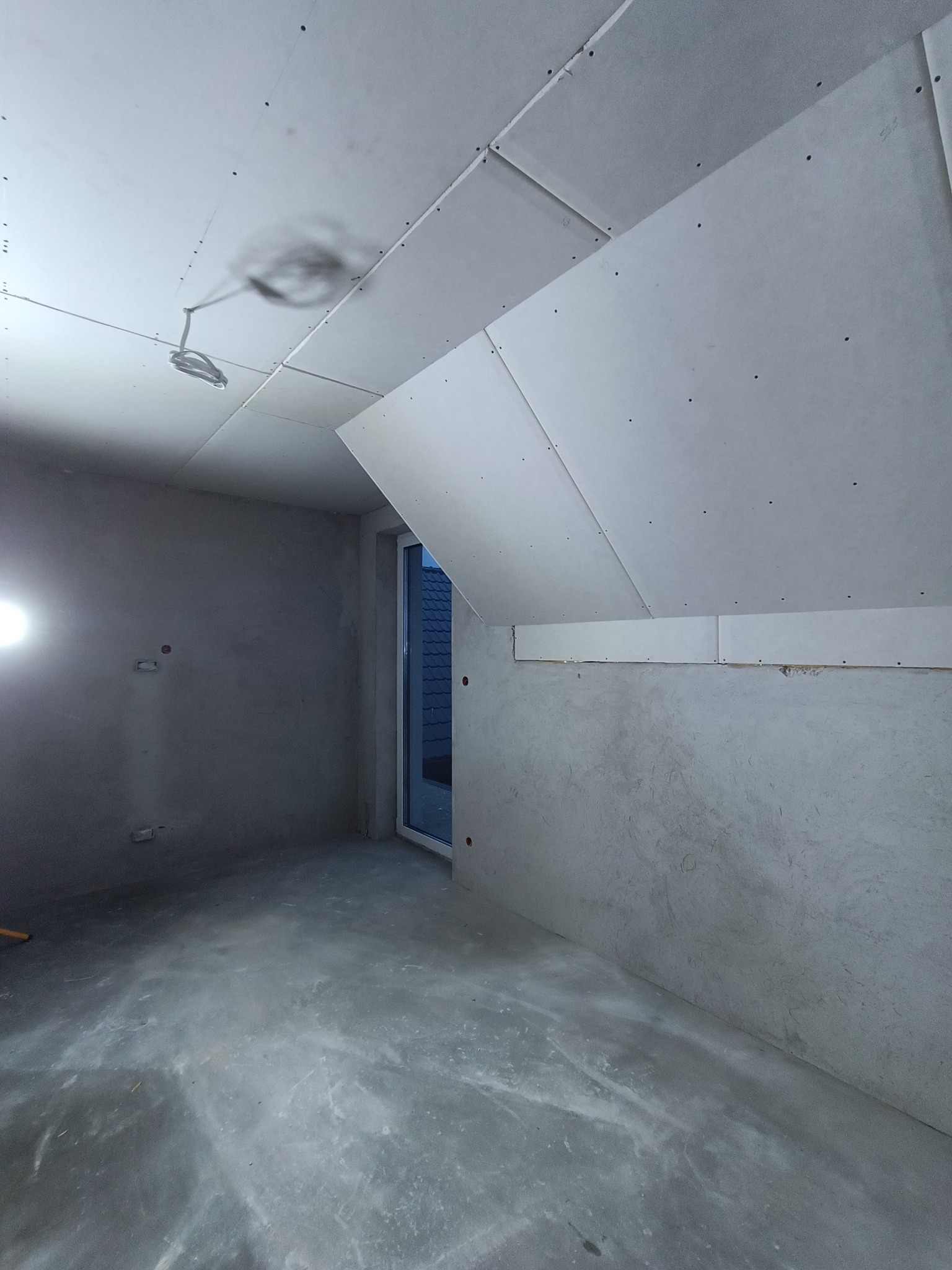 Wnętrze poddasza w trakcie wykańczania: surowe ściany, częściowo zamontowane płyty gipsowo-kartonowe na skosach sufitu, widoczne przewody elektryczne i niedokończona wylewka podłogowa.