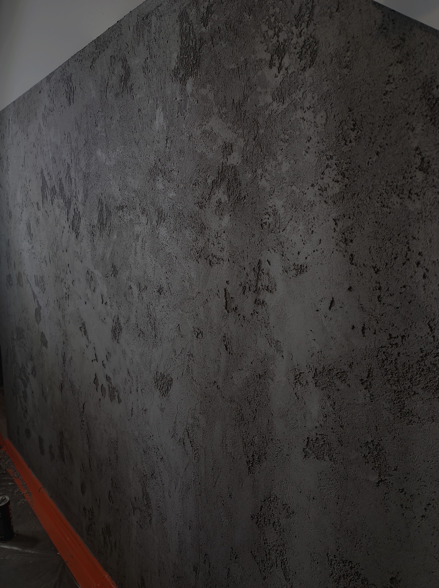 Ściana pokryta szarą, chropowatą strukturą, przypominającą beton architektoniczny. Widoczne nierówności i zagłębienia tworzące unikalny wzór. U dołu kadru, przy ścianie, widoczny pomarańczowy...