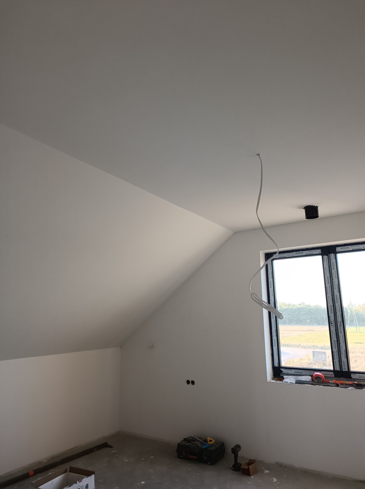 Poddasze po świeżym malowaniu na biało, z widocznym oknem w czarnej ramie, wystającym kablem ze sufitu i narzędziami na podłodze.