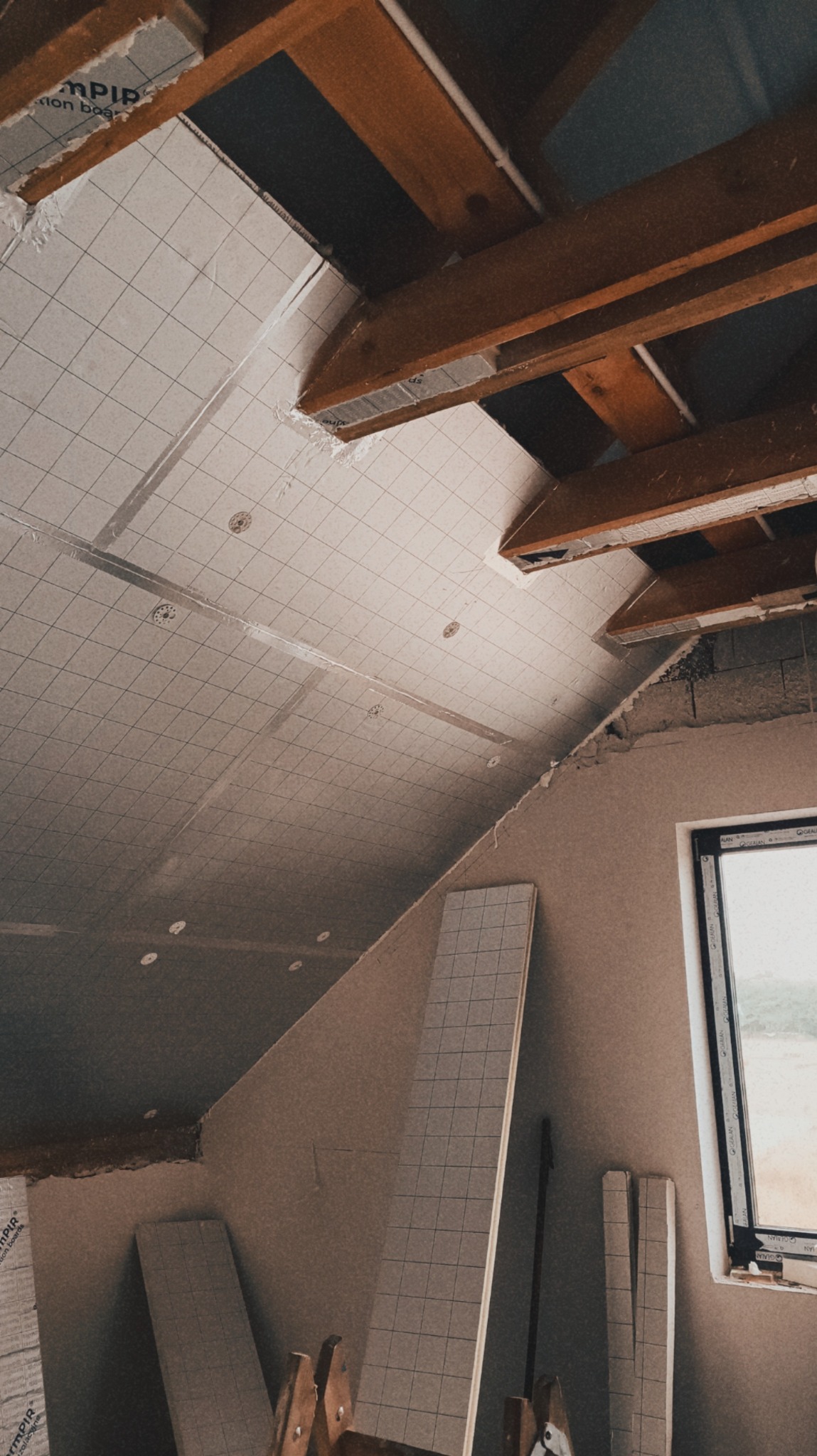 Poddasze w trakcie ocieplania: widoczne drewniane belki stropowe, płyty izolacyjne z nadrukowaną siatką, częściowo wykończona ściana szczytowa z oknem.