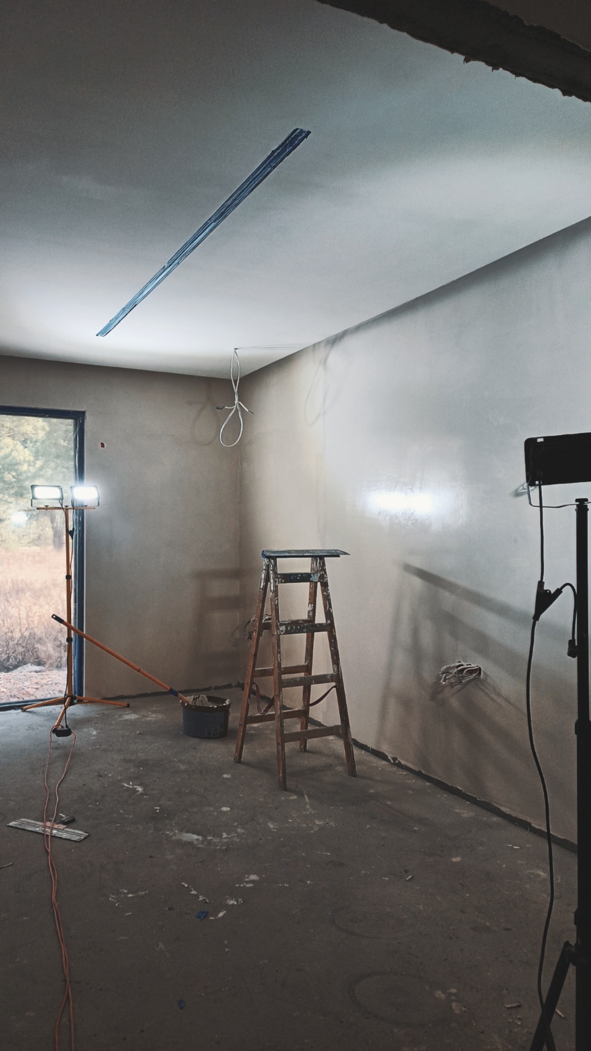 Wnętrze pokoju w trakcie prac wykończeniowych; ściany pokryte świeżo nałożoną warstwą, widoczna drewniana drabina, lampy budowlane, wiązka przewodów zwisająca z sufitu i listwa montażowa na suficie.