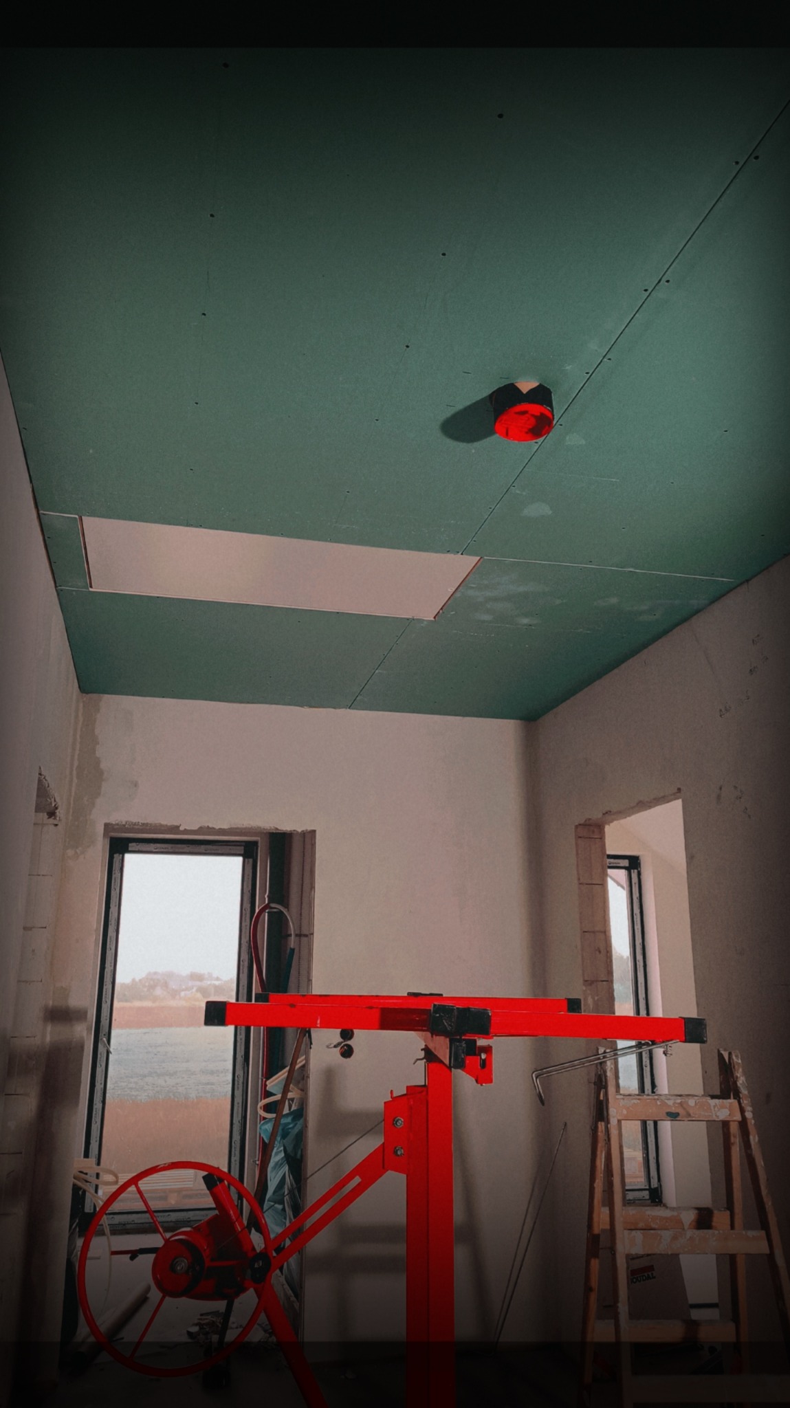Montaż zielonej płyty gipsowo-kartonowej na suficie z widocznym otworem na oświetlenie i czerwonym podnośnikiem do płyt.