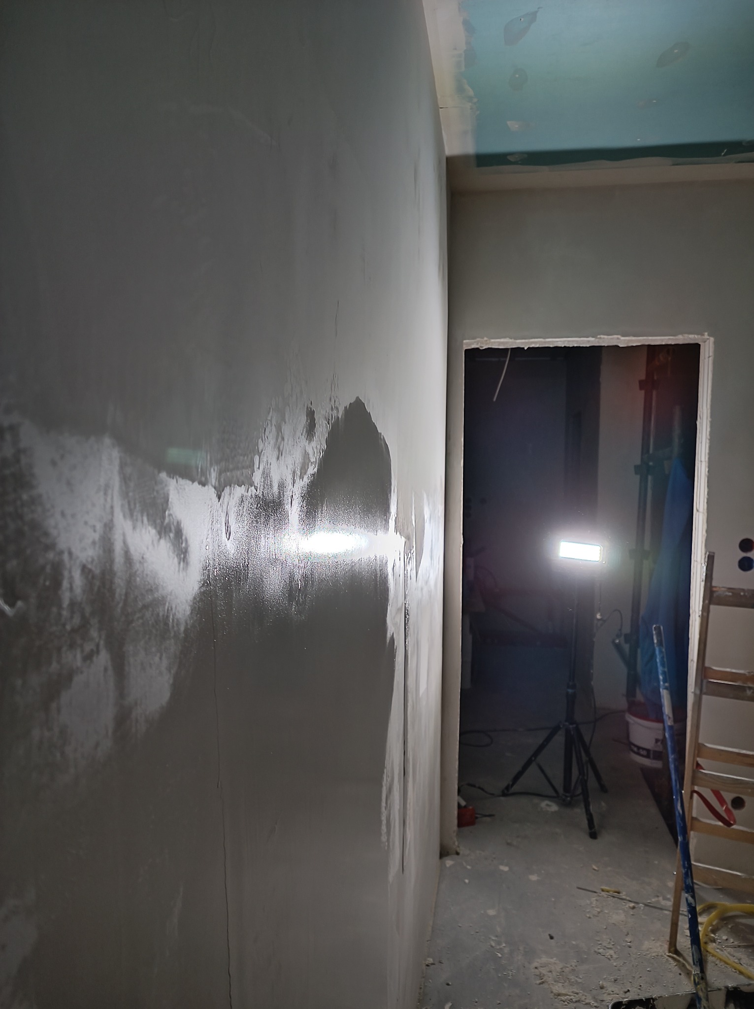 Świeżo nałożona warstwa mokrej, szarej masy na ścianie w trakcie prac wykończeniowych, widoczny fragment sufitu, drabina i lampa budowlana w tle.