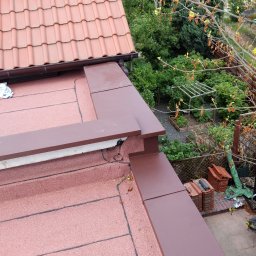 M-Tech - Widok z góry na fragment dachu z panelami słonecznymi, brązowe obróbki blacharskie i przygotowane dachówki, ogród w tle.