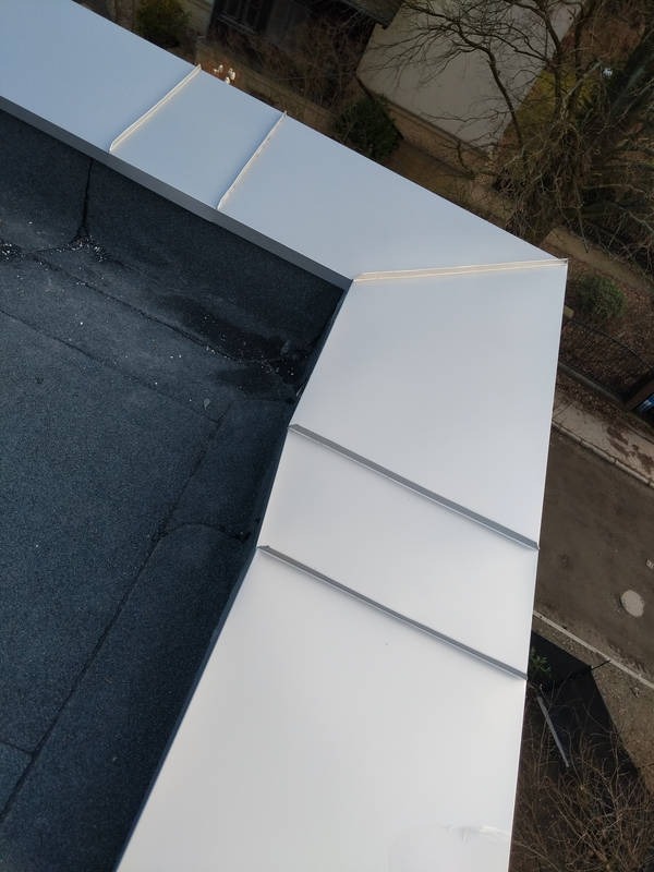 Szczegółowe ujęcie dachu pokrytego papą z nowymi, błyszczącymi obróbkami blacharskimi w kolorze srebrnym, widoczne z góry z elementami otoczenia.