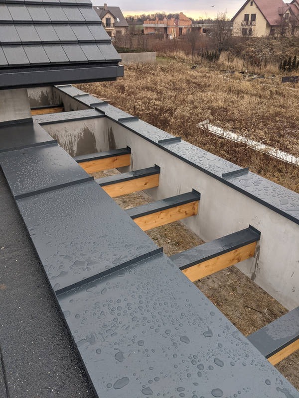 Szare, metalowe elementy konstrukcji dachu z widocznymi kroplami deszczu, drewniane belki wspierające, w tle zabudowa jednorodzinna i pole.