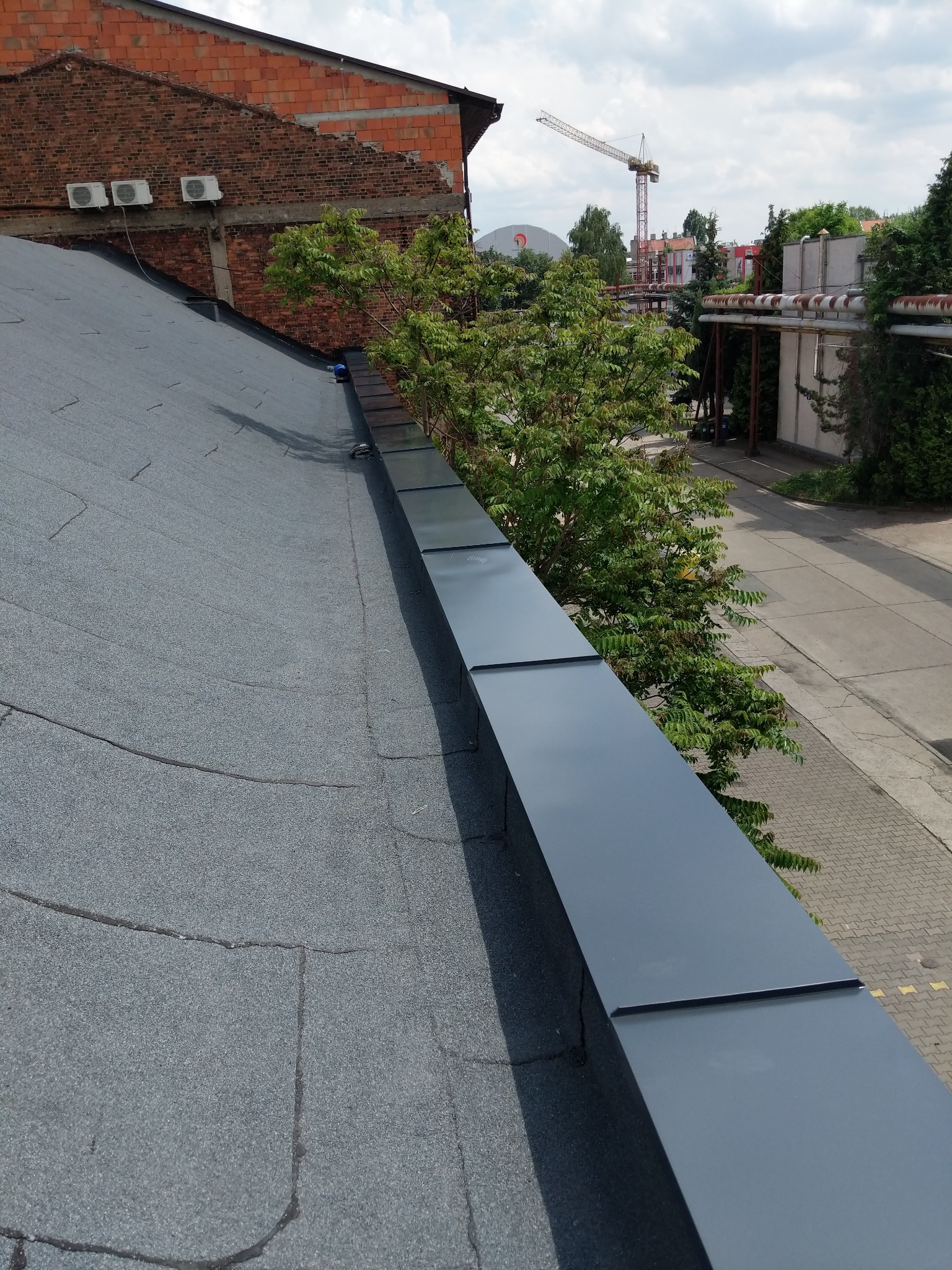 Widok z góry na płaski dach pokryty papą, z nową, błyszczącą, ciemnoszarą obróbką blacharską przy krawędzi, na tle drzewa i budynku z cegły z klimatyzatorami.