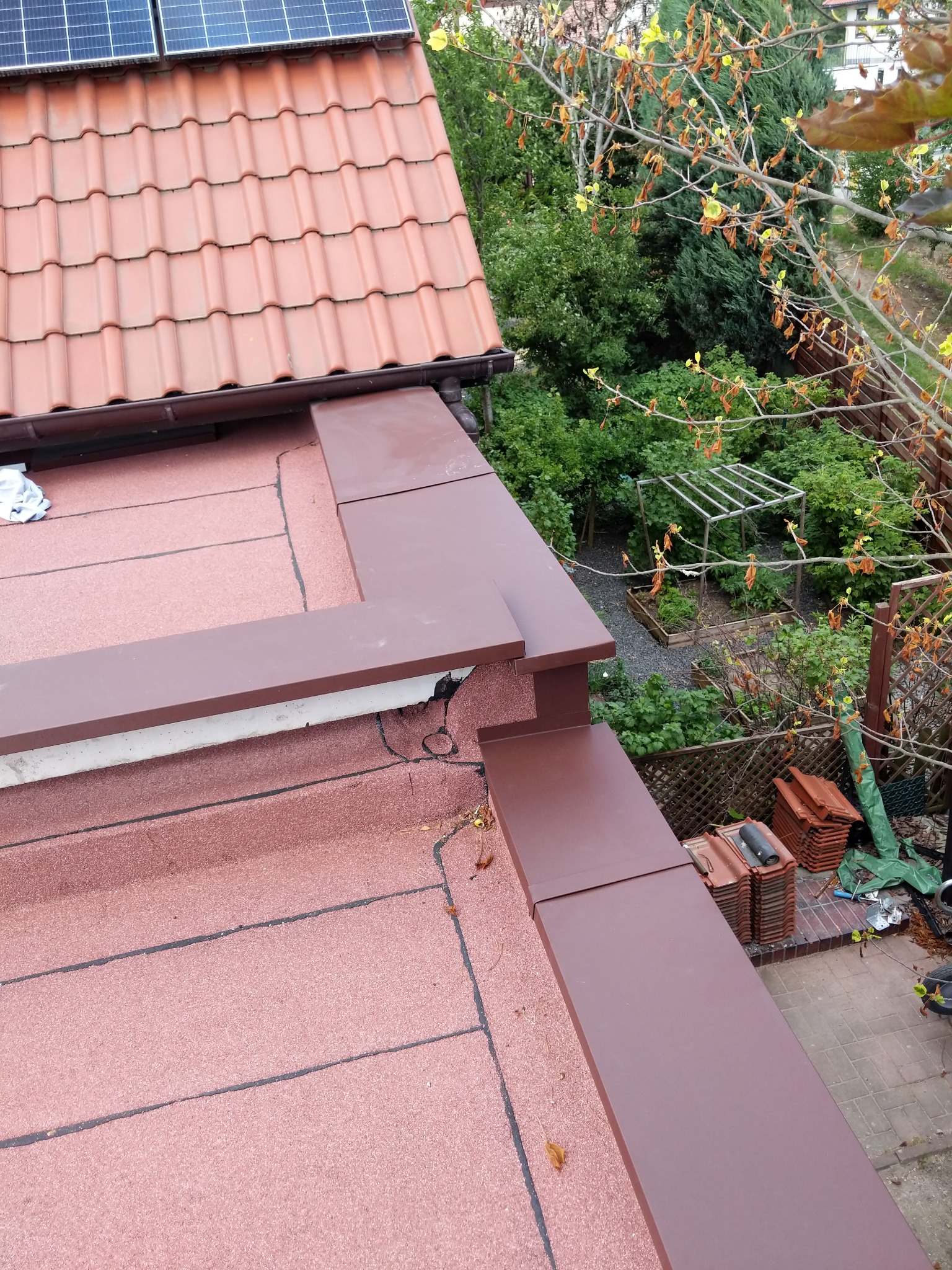 Widok z góry na fragment dachu z panelami słonecznymi, brązowe obróbki blacharskie i przygotowane dachówki, ogród w tle.