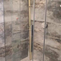 Firma remontowo budowlana - Szklana kabina prysznicowa z chromowanymi profilami, zamontowana w łazience z szarymi, marmurkowymi płytkami. Widoczna poziomica oparta o ścianę kabiny i niebieska taśma malarska na podłodze.