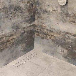 Firma remontowo budowlana - Nowoczesna kabina prysznicowa z szarymi płytkami imitującymi beton, odpływ liniowy w podłodze, bateria Grohe na ścianie.
