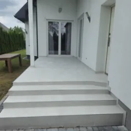 Wyłożone jasnoszarą płytką schody wejściowe do domu z tarasem, widoczna kostka brukowa i fragment ogrodu z tujami.