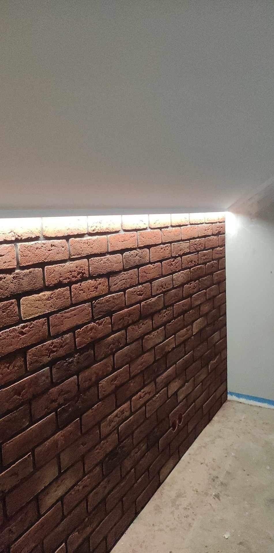 Ściana z cegieł klinkierowych z podświetleniem LED u góry, surowy beton na podłodze, widoczne otwory na gniazdka elektryczne.