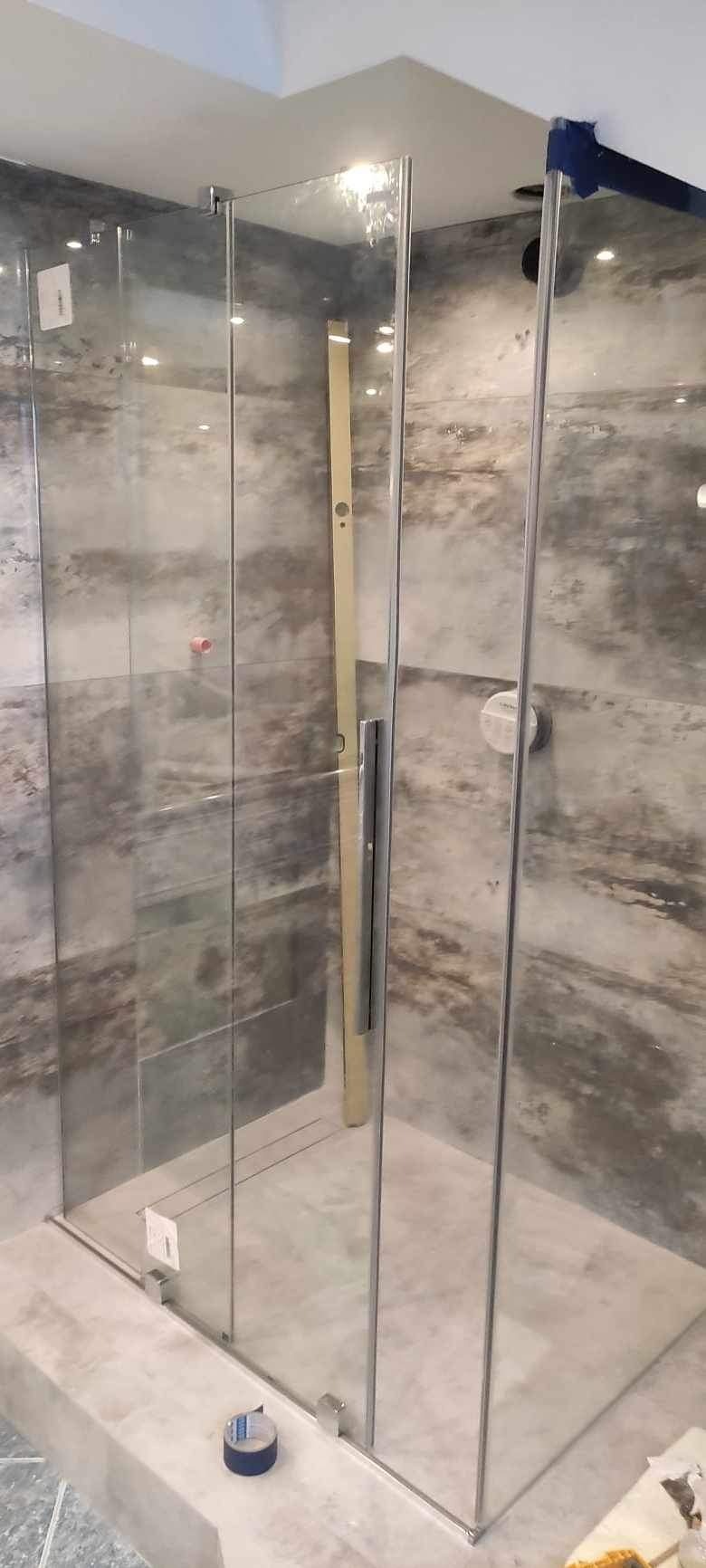 Szklana kabina prysznicowa z chromowanymi profilami, zamontowana w łazience z szarymi, marmurkowymi płytkami. Widoczna poziomica oparta o ścianę kabiny i niebieska taśma malarska na podłodze.