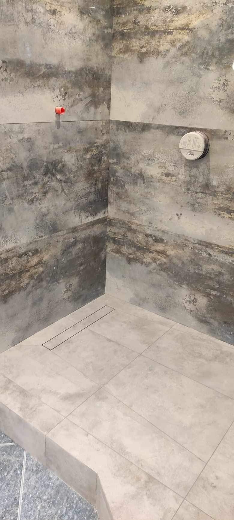 Nowoczesna kabina prysznicowa z szarymi płytkami imitującymi beton, odpływ liniowy w podłodze, bateria Grohe na ścianie.