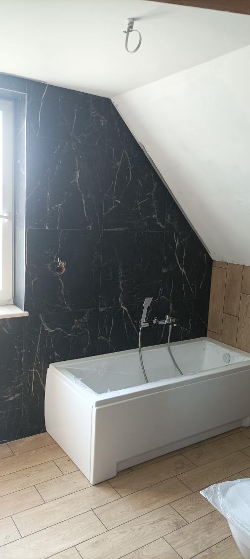 Wykończona łazienka z wanną, czarnymi płytkami imitującymi marmur na ścianie i drewnopodobną podłogą, widoczny uchwyt na słuchawkę prysznicową i instalacja elektryczna pod oświetlenie na suficie.