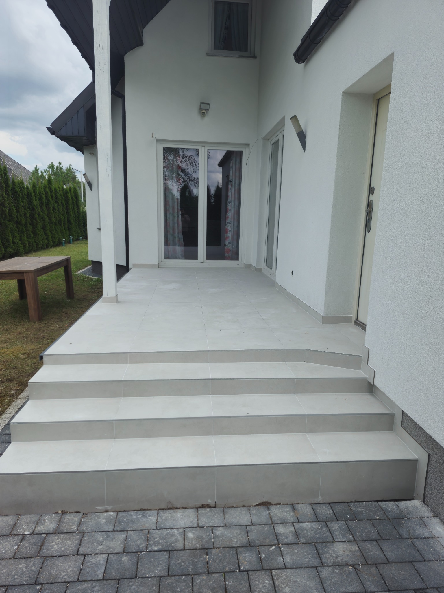 Wyłożone jasnoszarą płytką schody wejściowe do domu z tarasem, widoczna kostka brukowa i fragment ogrodu z tujami.