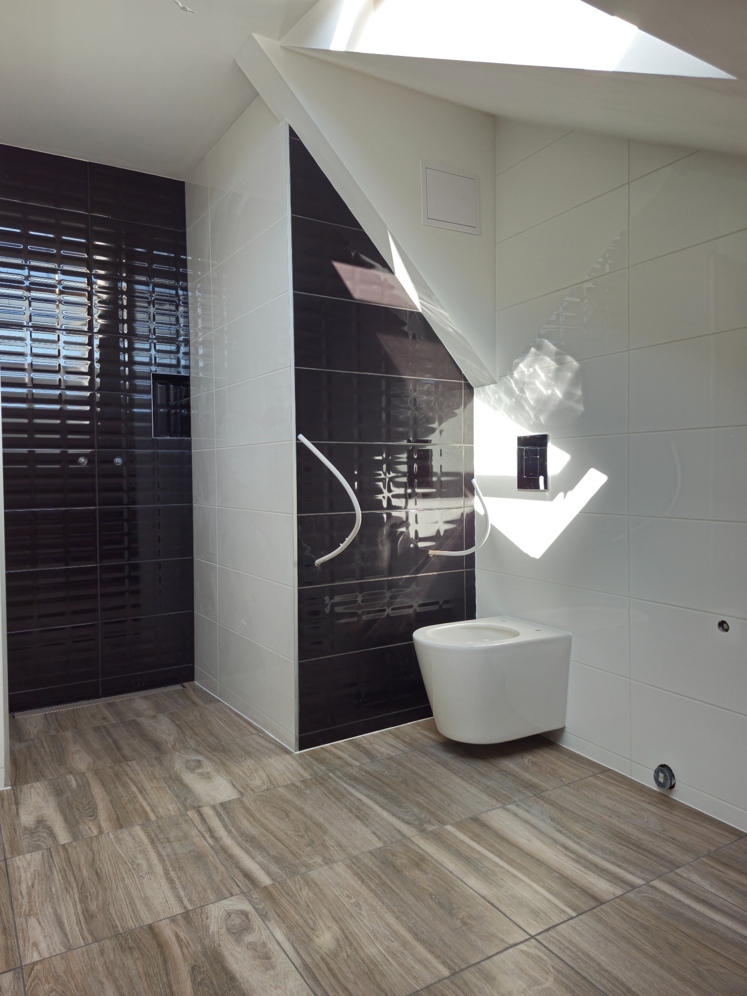 Nowoczesna łazienka na poddaszu z czarno-białymi płytkami ściennymi i podłogą imitującą drewno, widoczne przyłącza do armatury.