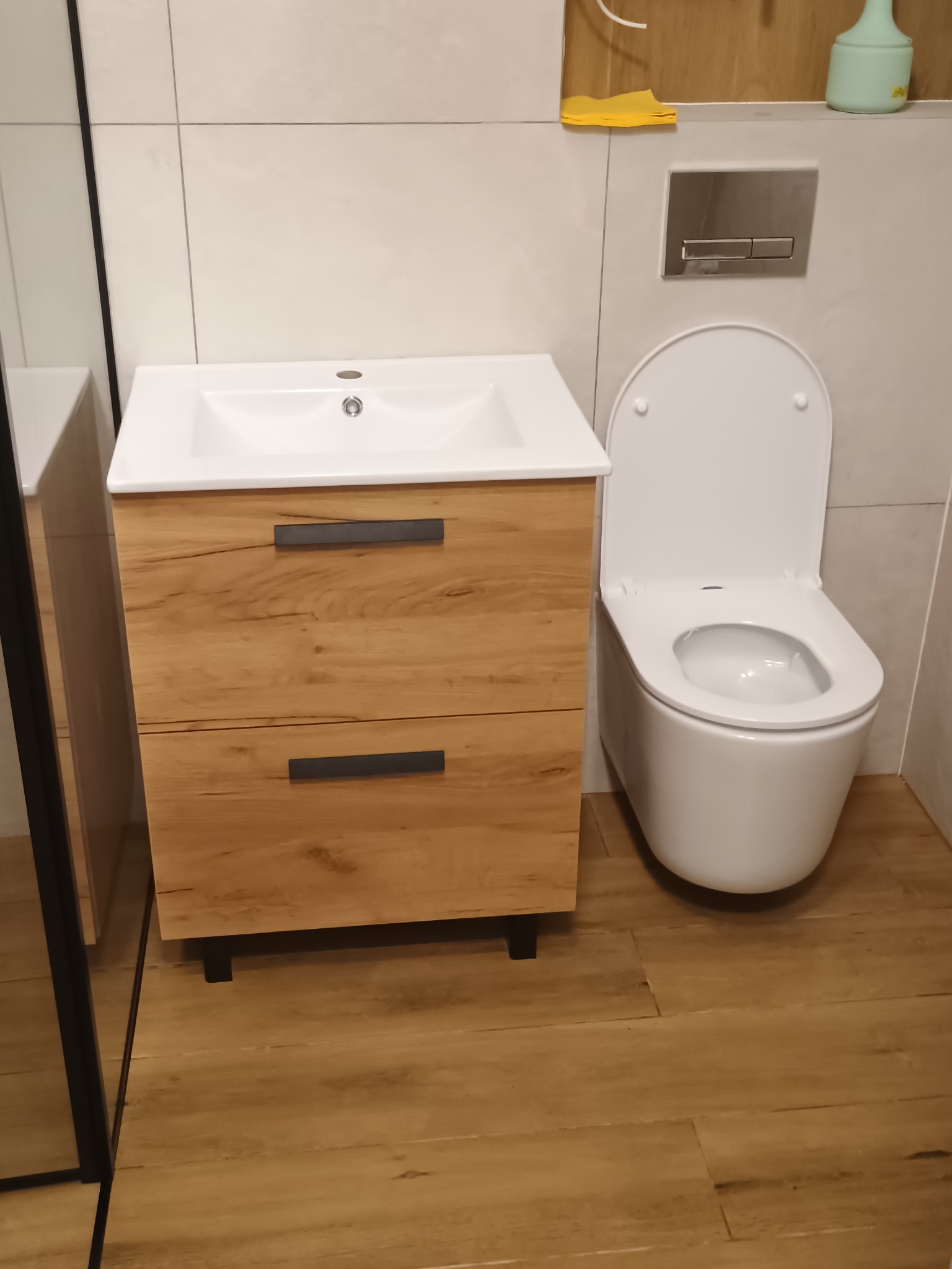 Nowoczesna łazienka z drewnianą szafką pod umywalką i podwieszaną toaletą. Jasne płytki na ścianach i podłodze. Minimalistyczny design, czyste linie.