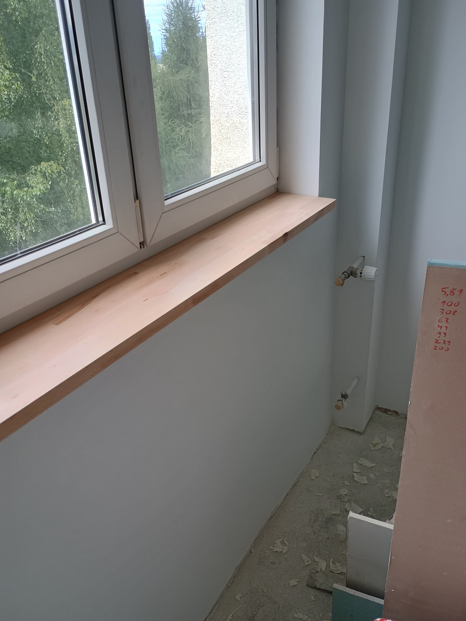 Jasny parapet z drewna w oknie, świeżo pomalowane ściany na szaro, widoczne rury grzewcze i przygotowane płyty. Wnętrze w trakcie prac wykończeniowych.