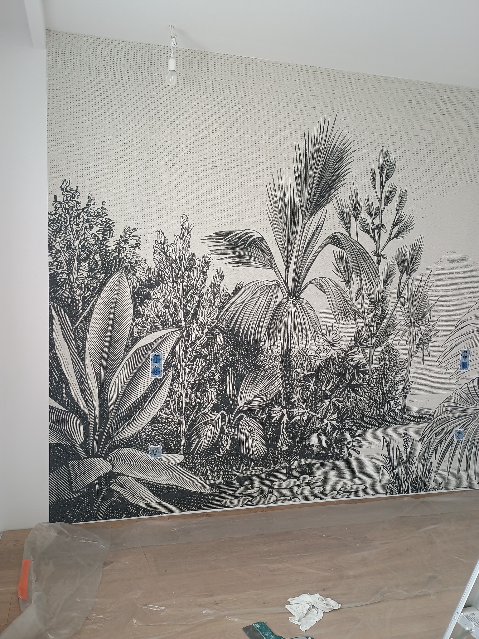 Tapeta w stylu retro z motywem tropikalnej roślinności na ścianie, zabezpieczona podłoga folią malarską, widoczne gniazdka elektryczne.