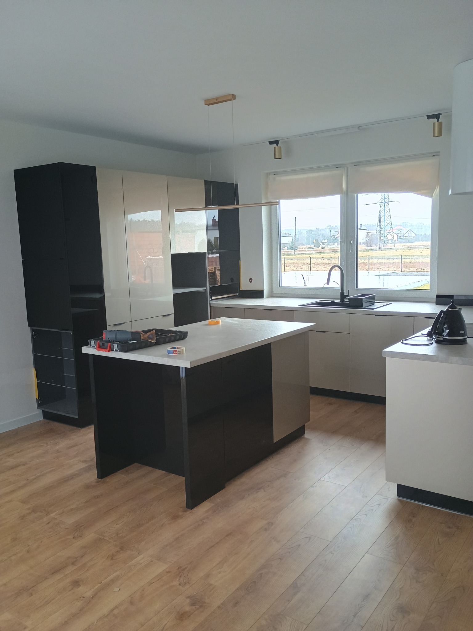 Nowoczesna kuchnia w trakcie montażu: wyspa kuchenna z narzędziami, jasne szafki, okno z widokiem na zewnątrz. Widoczne elementy wykończenia i oświetlenie.