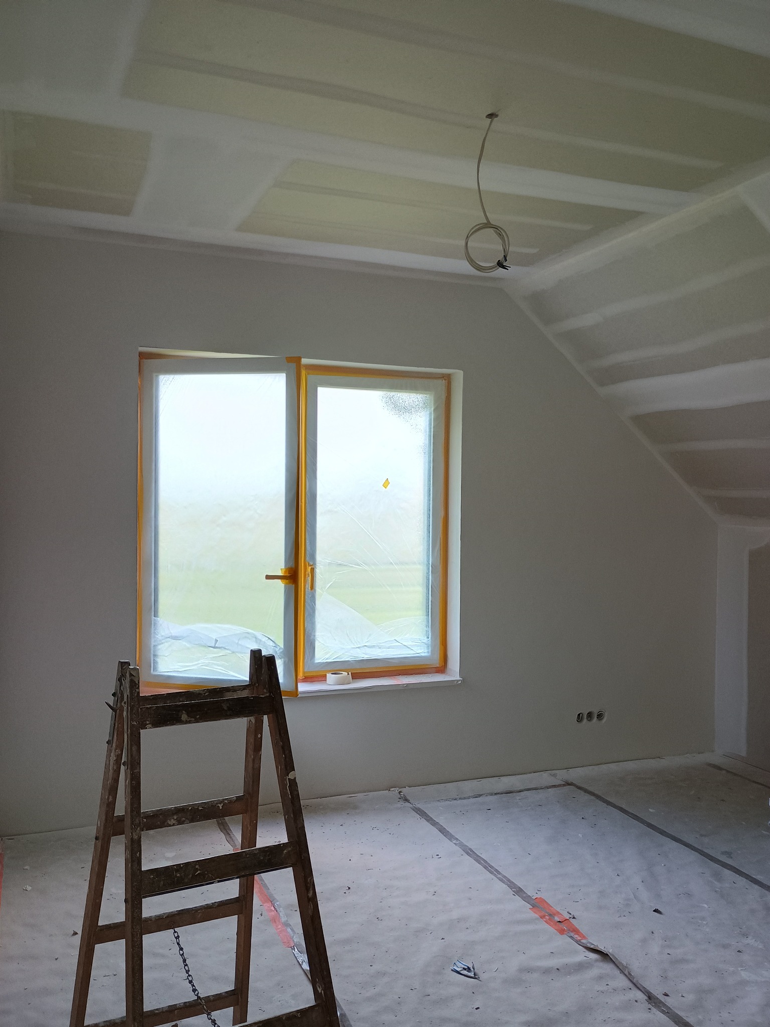 Pokój w trakcie prac wykończeniowych: ściany po szpachlowaniu, zabezpieczone okno folią, drewniana drabina, widoczne gniazdka elektryczne i przewód oświetleniowy.