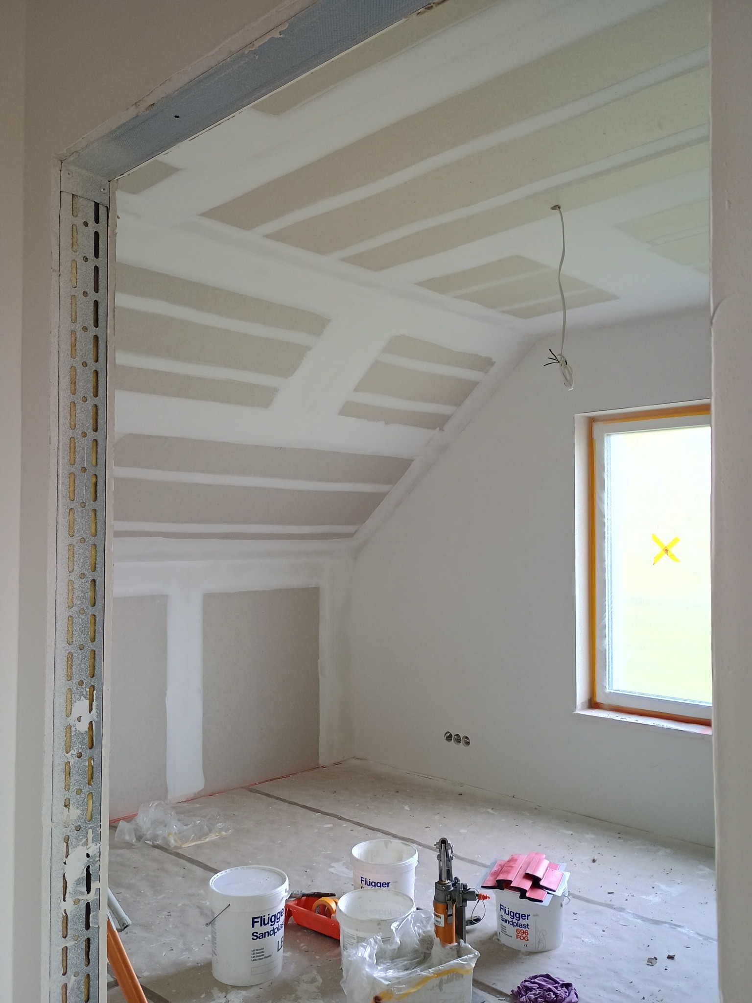 Poddasze w trakcie prac wykończeniowych: widoczne płyty karton-gips na suficie, metalowe profile przy otworze drzwiowym, okno z taśmą zabezpieczającą i farby na podłodze.