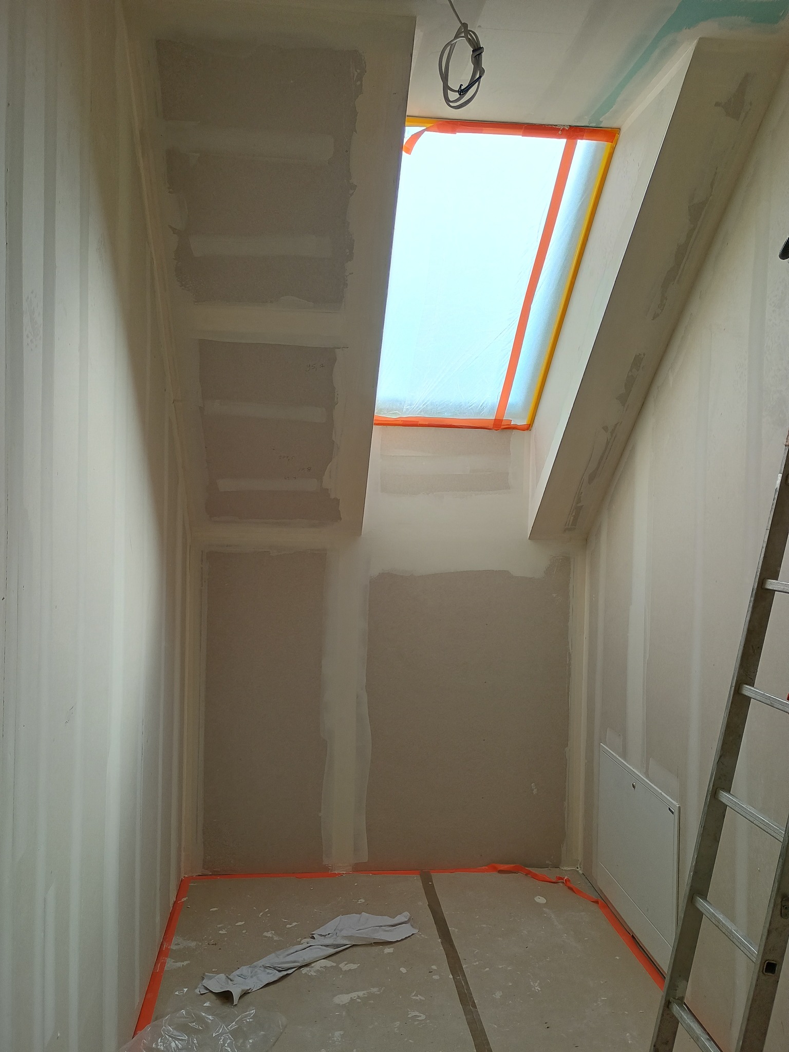 Poddasze w trakcie prac wykończeniowych: ściany z płyt gipsowych, zabezpieczone okno dachowe, widoczna drabina i folia ochronna na podłodze. Etap szpachlowania.