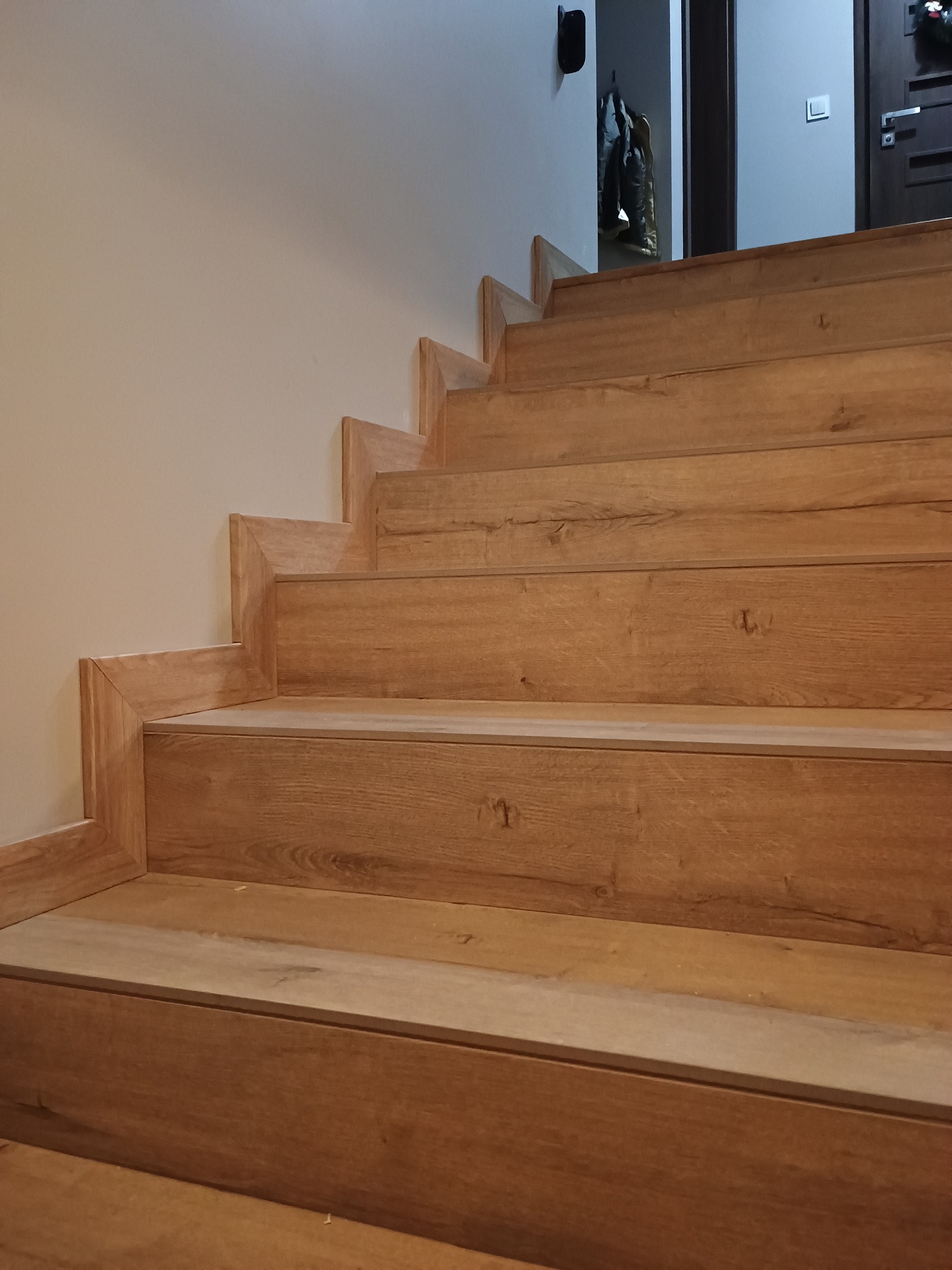 Wykończone schody z drewnopodobnych paneli, widok z dołu. Jasne drewno, minimalistyczny design, fragment ściany i drzwi w tle. Detal wykończenia wnętrza.