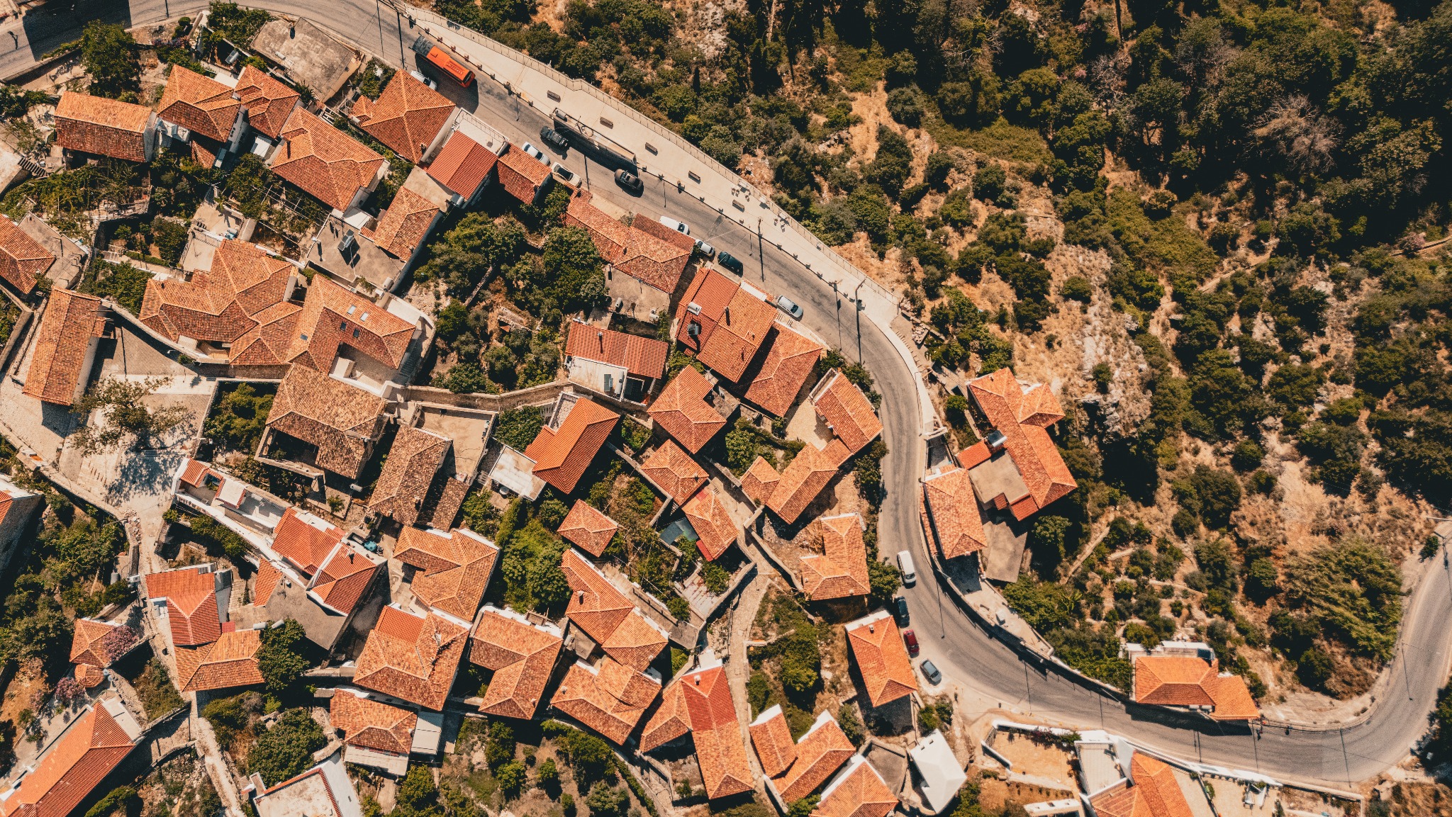 Widok z lotu ptaka na miasteczko z domami o czerwonych dachach, otoczone zielenią i krętą drogą z samochodami.