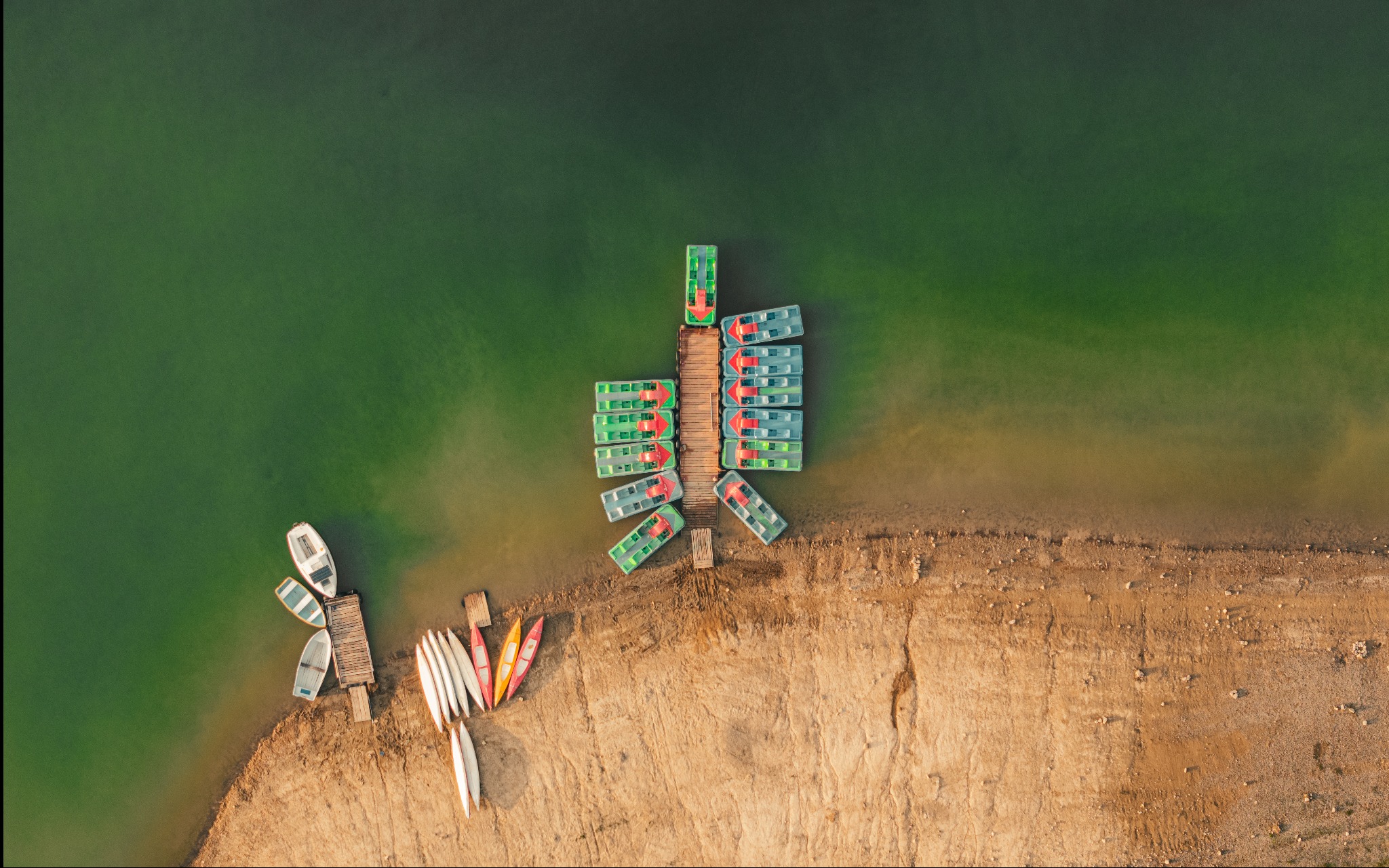 Widok z góry na przystań z łódkami i kajakami na brzegu jeziora o zielonkawej wodzie, w tle piaszczysta plaża.