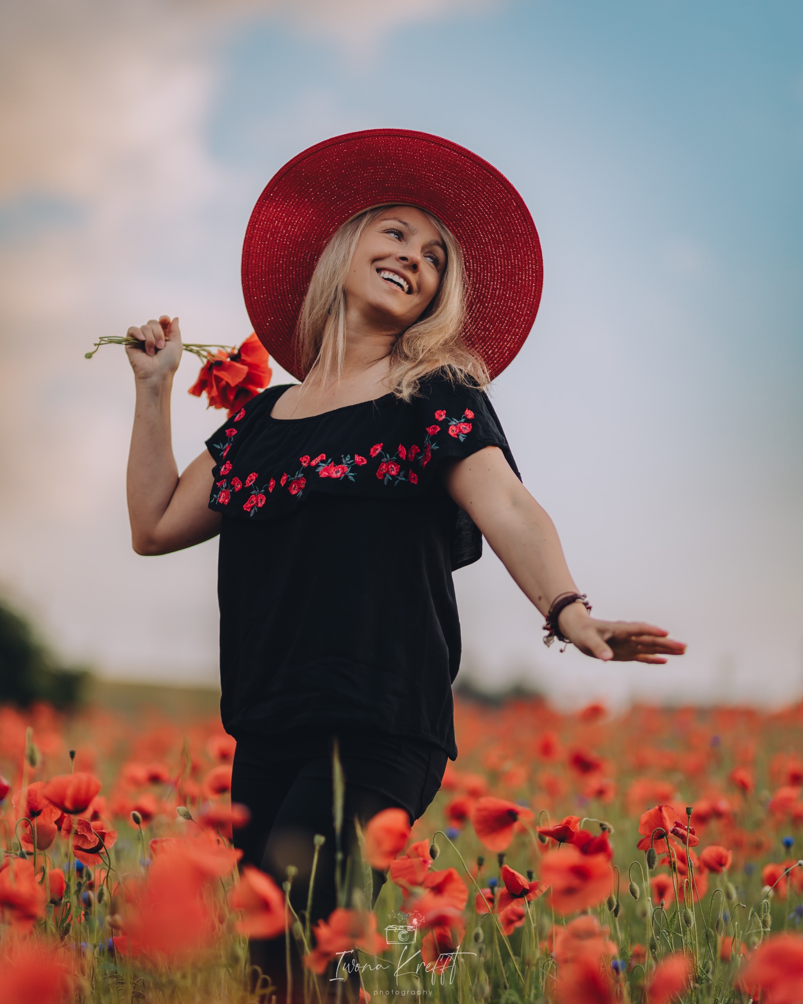 Uśmiechnięta blondynka w czarnej bluzce z haftowanymi czerwonymi kwiatami i czerwonym kapeluszu na polu maków, trzymająca bukiet maków w dłoni.