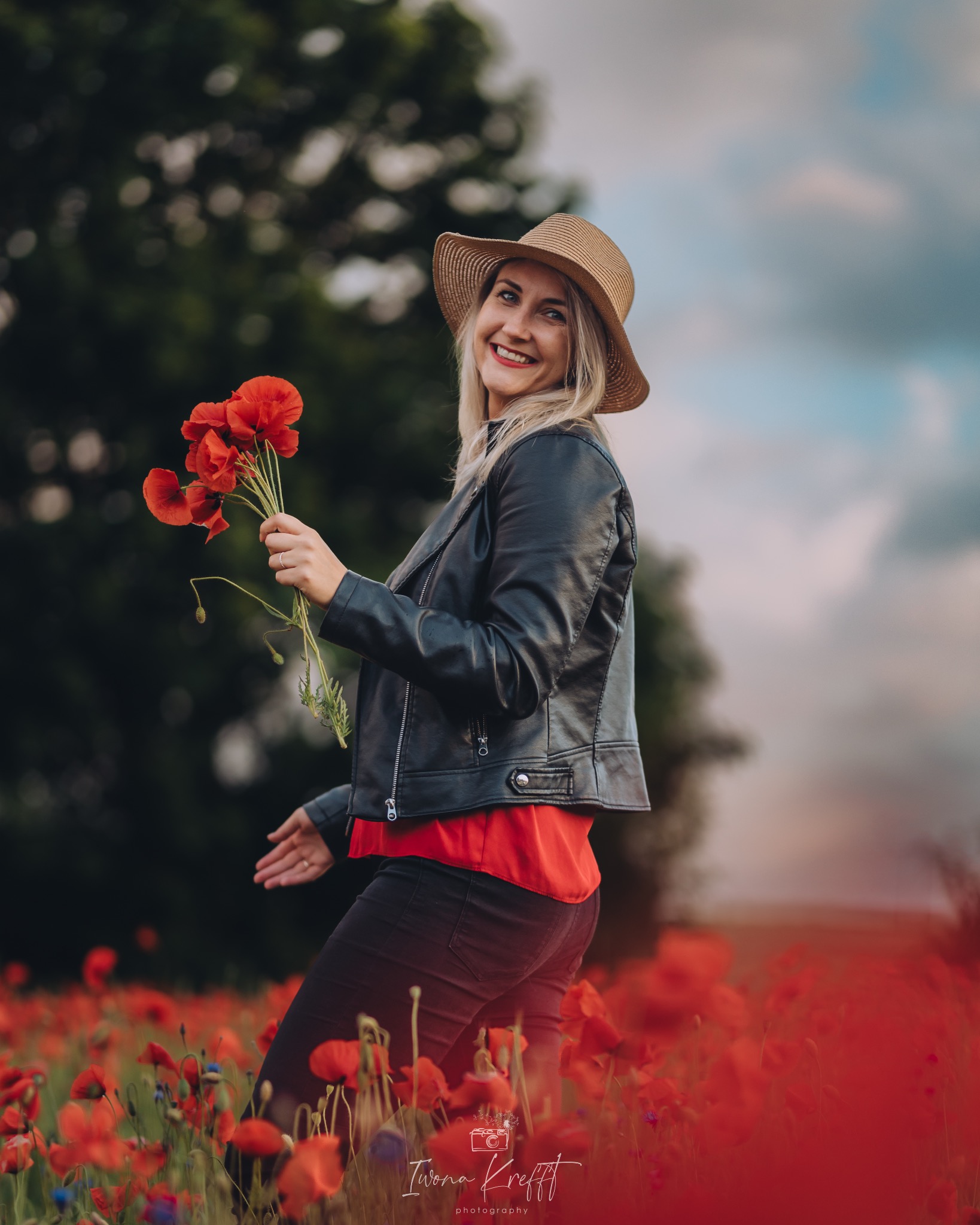 Uśmiechnięta blondynka w kapeluszu i czarnej kurtce trzyma bukiet czerwonych maków na tle pola makowego i zachmurzonego nieba.