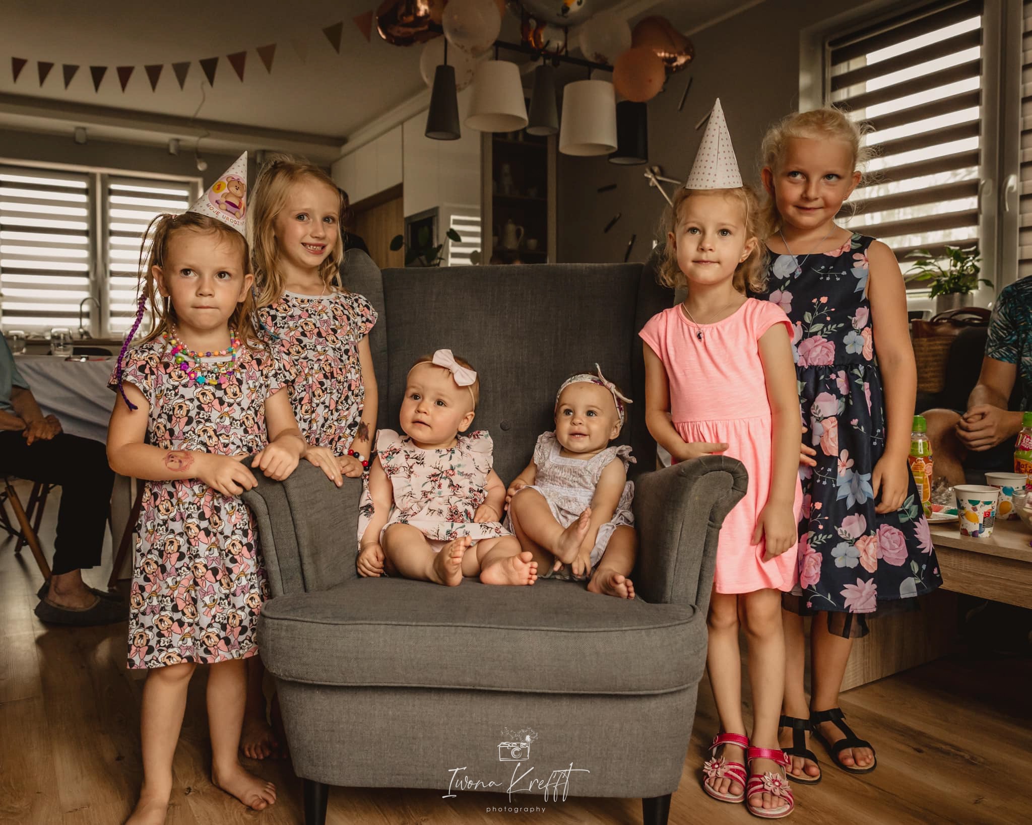 Grupa dzieci w różnym wieku, w tym dwie małe dziewczynki siedzące w szarym fotelu, pozuje do zdjęcia w pomieszczeniu udekorowanym balonami i girlandami.