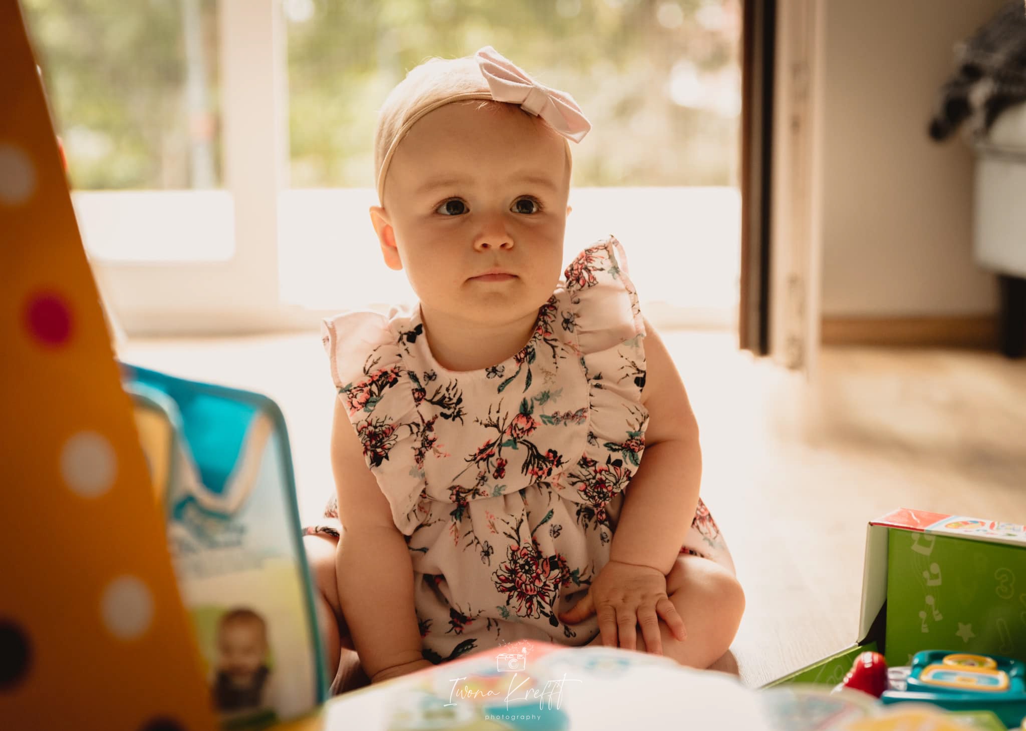 Urocza dziewczynka z kokardą na głowie siedzi na podłodze w jasnym pokoju, otoczona zabawkami, ubrana w sukienkę w kwiaty z falbankami.