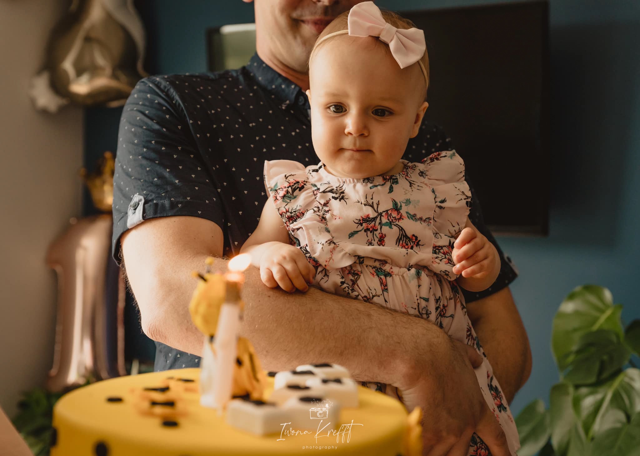 Uśmiechnięty tata trzyma na rękach roczną córeczkę z różową kokardką na głowie, przed nimi żółty tort urodzinowy ze świeczką w kształcie żyrafy.