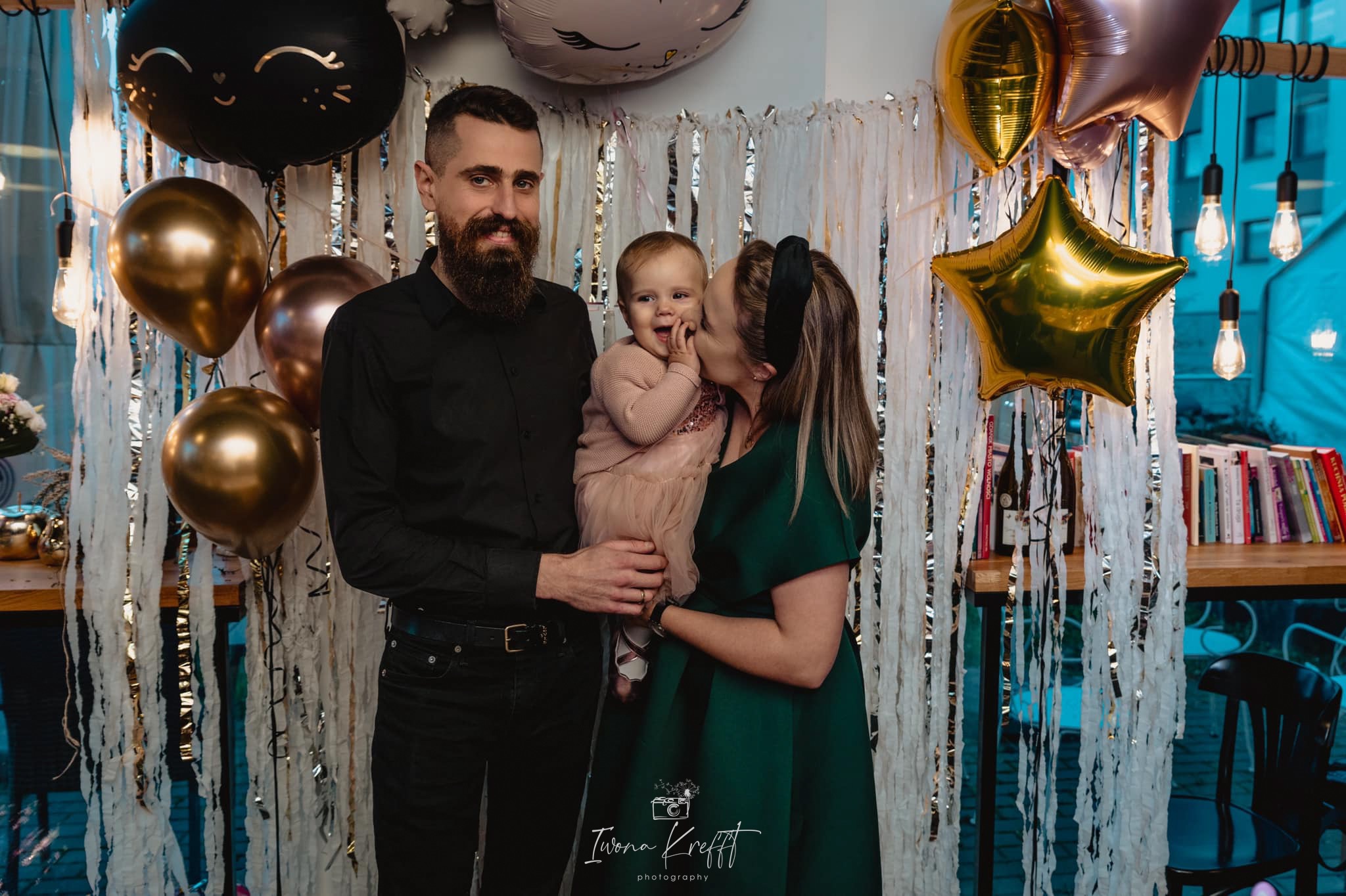 Rodzinne zdjęcie z małą dziewczynką w różowej sukience, mama całuje córkę w policzek, tata stoi obok na tle dekoracji z balonów i białych frędzli.