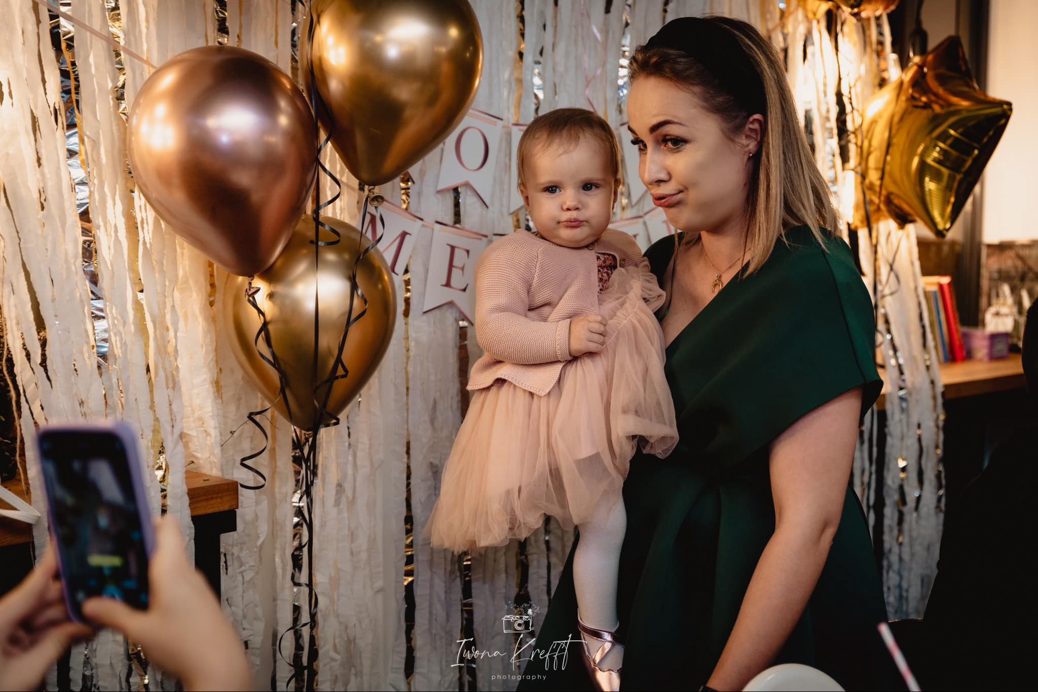 Matka trzymająca małą dziewczynkę w różowej sukience na tle dekoracji urodzinowych z balonami i banerem 'ONE', fotografowana telefonem.